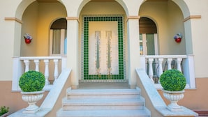 A building entrance with a green tiled door, white balustrades, and two potted topiary plants.