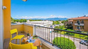 A balcony with yellow chairs, a table with a bottle and grapes, and a view of a street and buildings. - Casa Michelangelo Lake Garda Terrace Apt Sleeps 6 + AC (Desenzano del Garda)