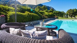 Un área junto a la piscina con mobiliario de mimbre, una sombrilla y vistas a la montaña.