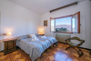 A bedroom with a bed, bedside lamps, a radiator, a window with a view, and a wooden chair. - Villa Giorgia Lake View Villa With Private Pool (Castelveccana)
