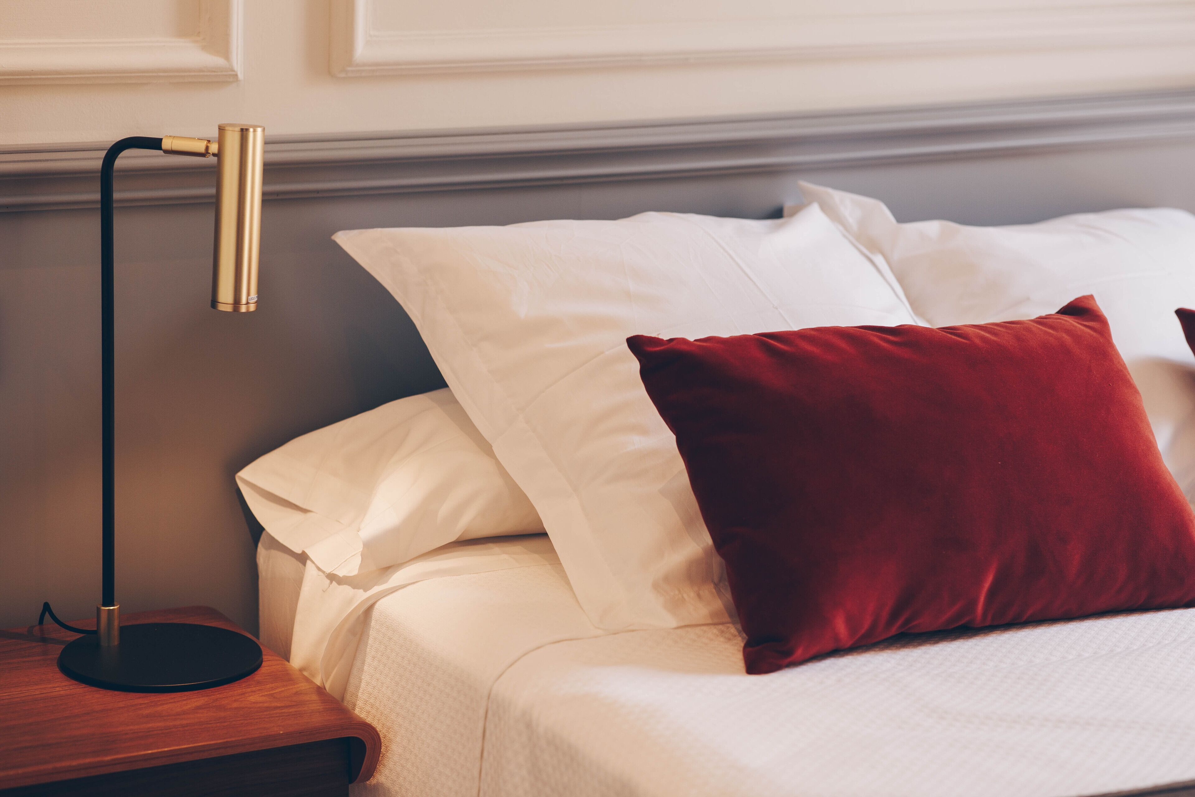 A neatly made bed with white linens and a red pillow, a black floor lamp, and a wooden nightstand.