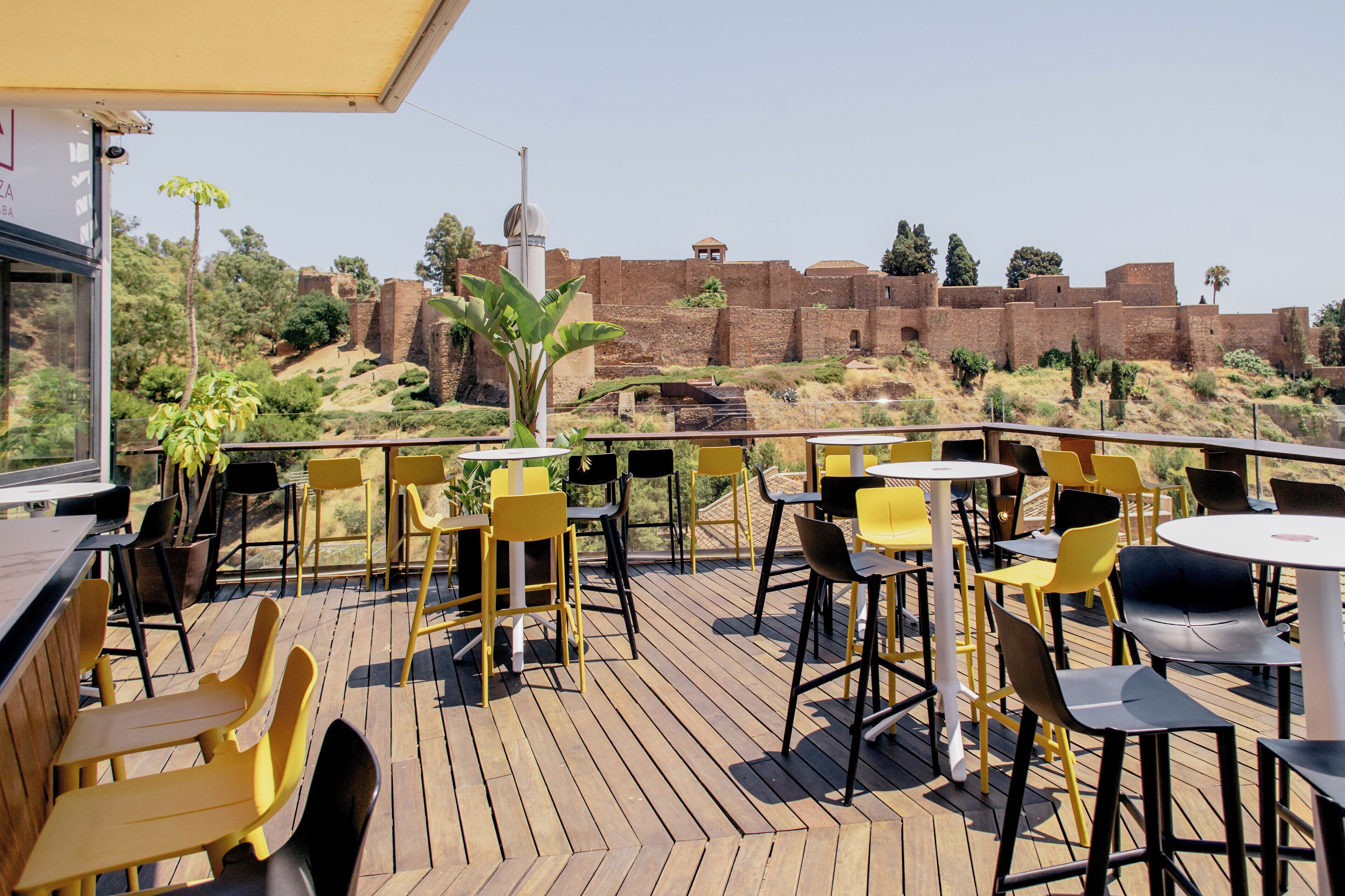 An outdoor patio with yellow and black chairs and tables, overlooking a historic brick wall and greenery.