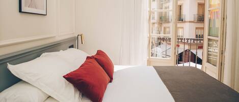 A hotel room with a bed, white and red pillows, a framed picture on the wall, and a view of a building with balconies.