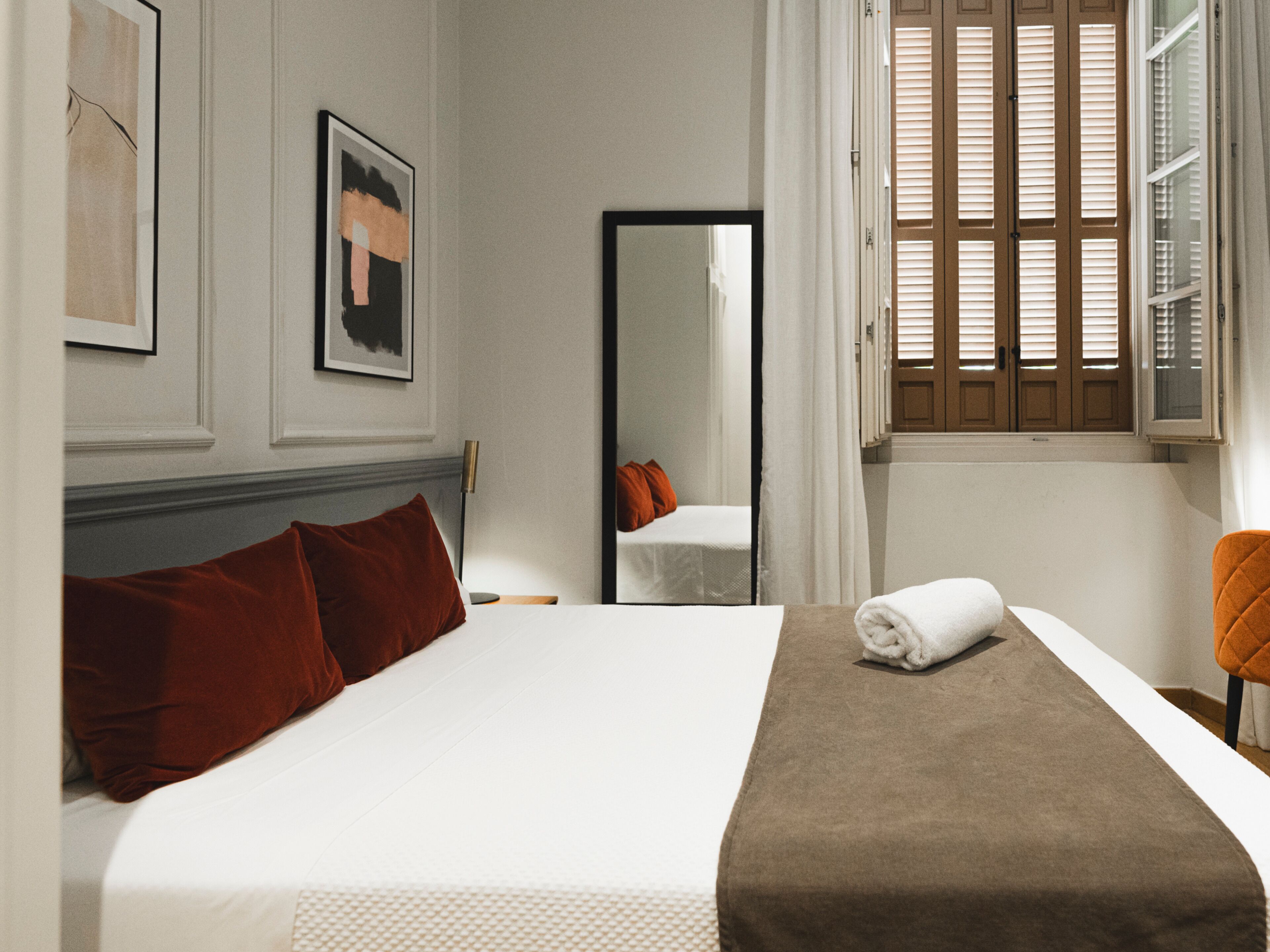 A neatly made bed with a white and brown bedspread, a red pillow, and a folded towel on the side.