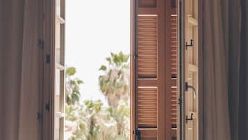 Ein Fenster mit geöffneten Holzläden, durch das sich Grünanlagen zeigen.