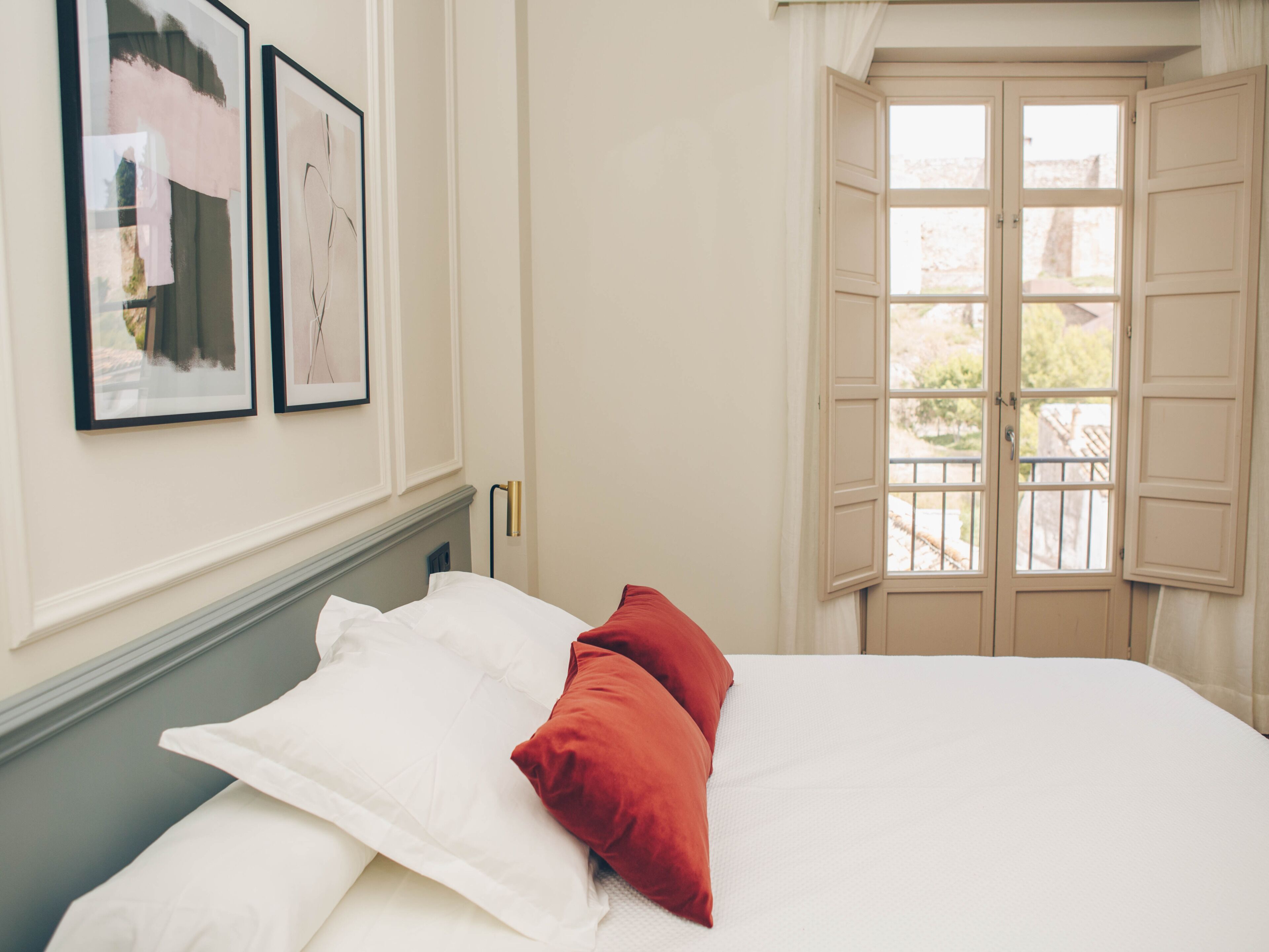 Ein ordentlich bezogenes Bett mit weißen und roten Kissen, ein gerahmtes Bild an der Wand und ein Fenster mit geöffneten Fensterläden.
