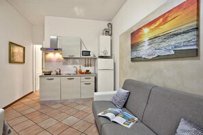 A compact living space with a kitchenette, a grey sofa with a magazine, and a painting of a sunset over the ocean. - Berny Salo: Near the Lake & Garage (Salò)