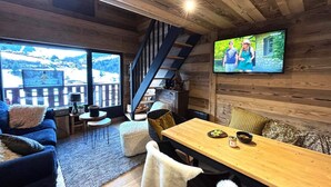 Una sala de estar acogedora con detalles de madera, un ventanal con vistas a un paisaje nevado, una escalera y un televisor en la pared.