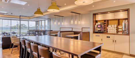 A modern restaurant with a long communal table, bar stools, pendant lighting, and a kitchen area with appliances.