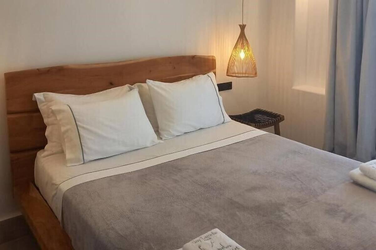 Un lit bien fait, avec du linge de lit blanc et une couverture grise, une tête de lit en bois, une lampe de chevet et une petite table de nuit.