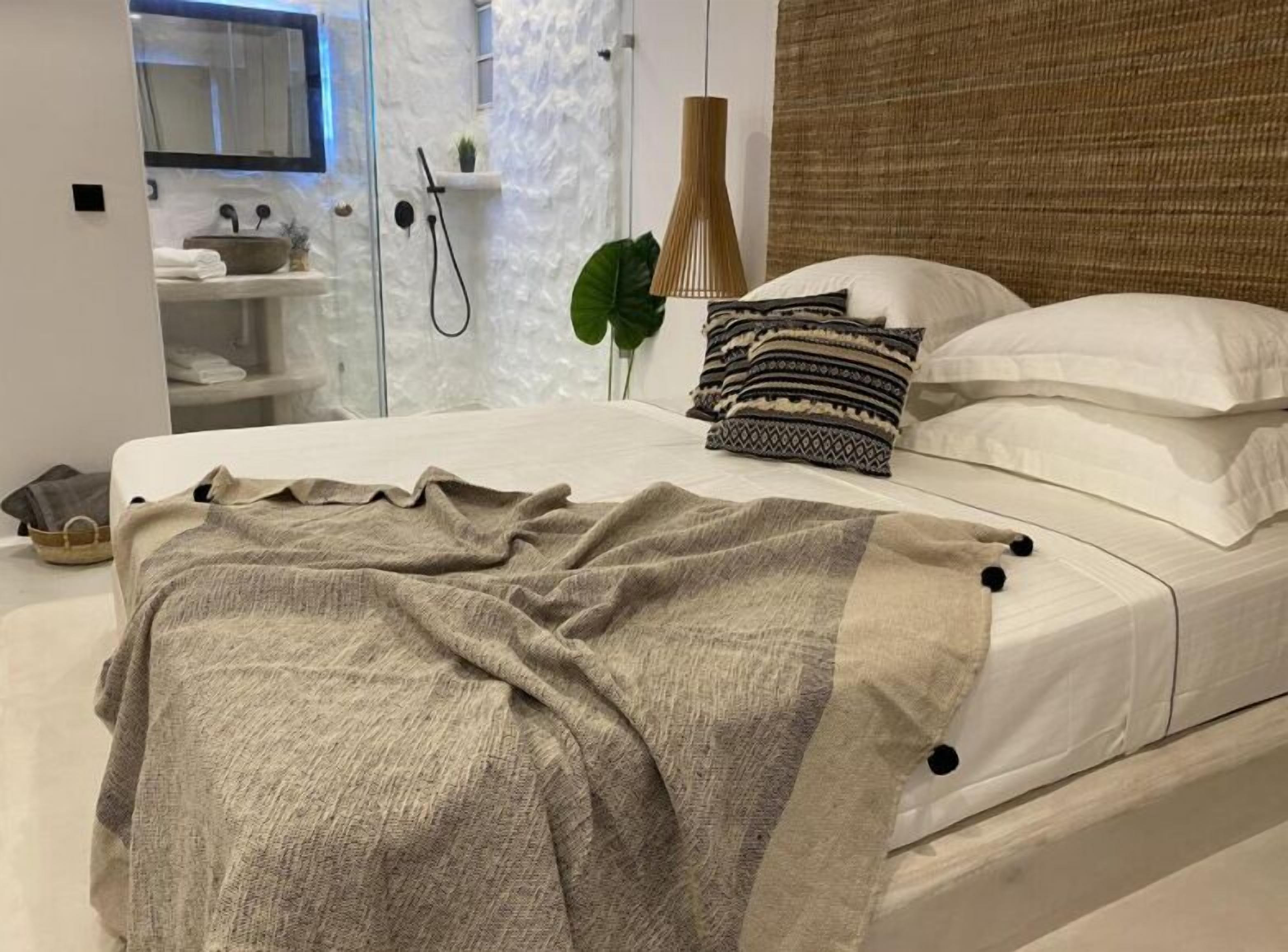 A modern bedroom with a large bed, a bedside table, a hanging lamp, and a view of a bathroom with a shower and a sink.