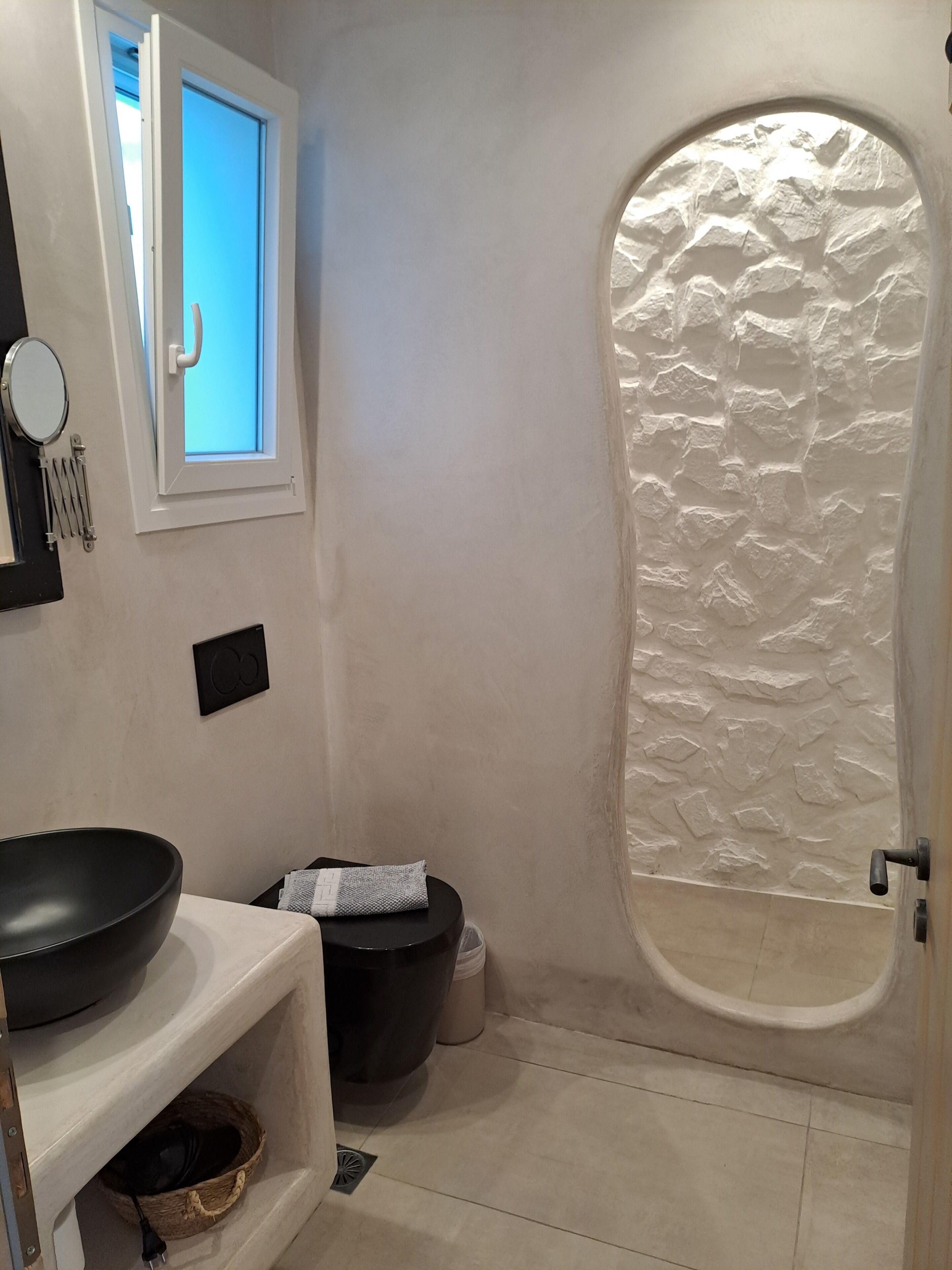 A bathroom with a stone-textured bathtub, a white sink with a black basin, a mirror, and a window with a blue view.