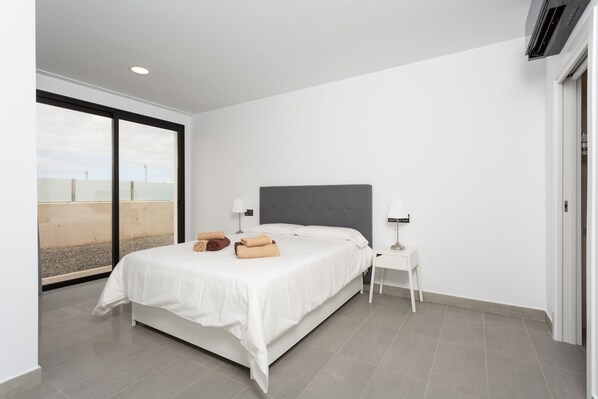 A modern bedroom with a large bed, two bedside tables with lamps, a grey upholstered headboard, and a view of the city through floor-to-ceiling windows.