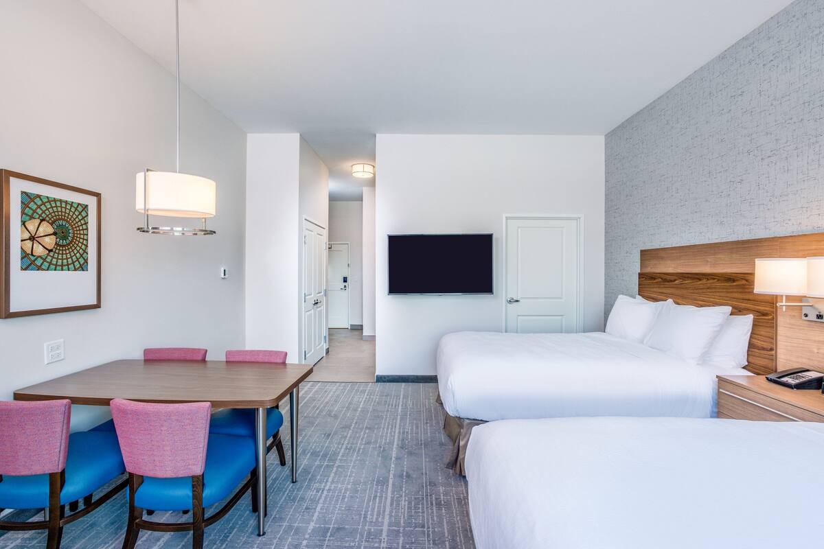 A hotel room with two beds, a dining area with chairs, a TV, and a wall-mounted phone.