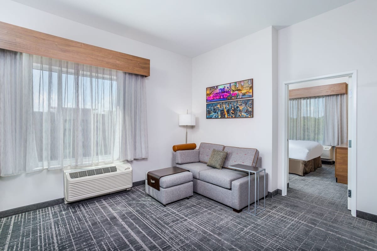 A hotel room with a grey sectional sofa, a small table, a lamp, a bed, a window with sheer curtains, a radiator, and a cityscape painting on the wall.