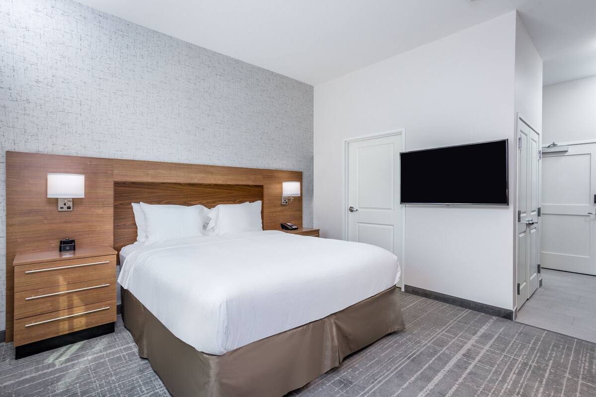 A hotel room with a large bed, a wooden headboard, bedside lamps, a TV mounted on the wall, a white door, and a small bedside table with drawers.