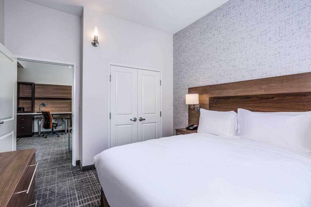 A hotel room with a large bed, a desk with a chair, a wardrobe, and a bathroom visible through an open door.