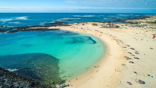 Ein Strand mit klarem türkisblauem Wasser, einem sandigen Ufer und einigen Gebäuden im Hintergrund.