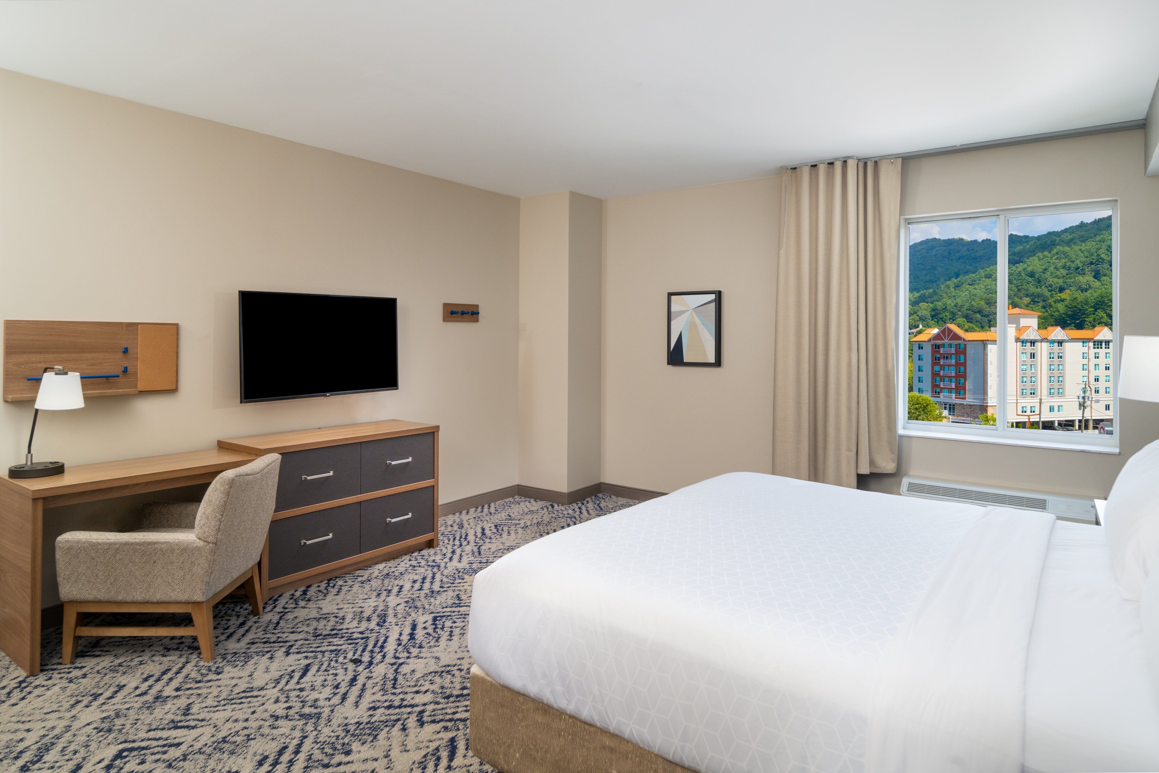 A hotel room with a bed, desk, chair, TV, and a window with a city view.