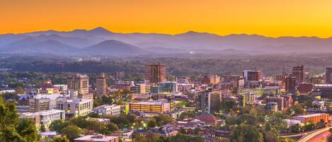 A cityscape with a variety of buildings, including a mix of modern and older architecture, set against a backdrop of mountains during sunset.