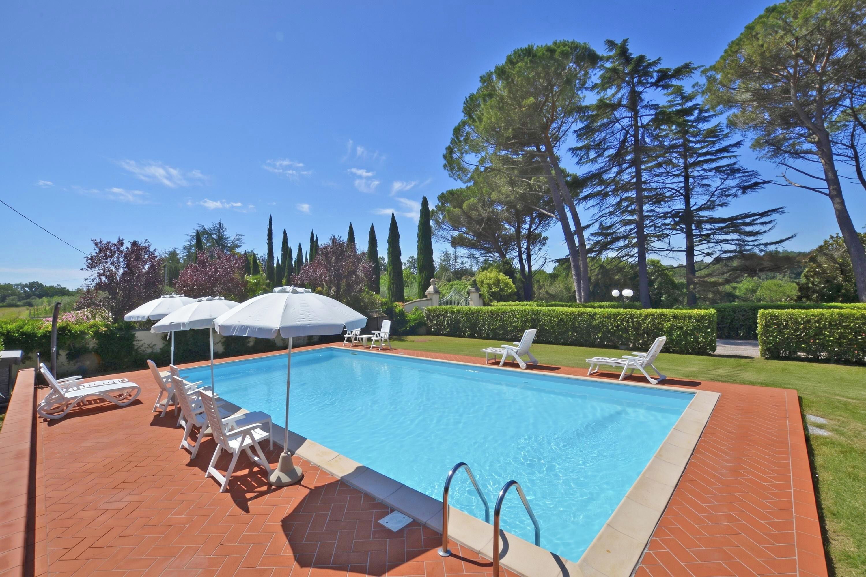 Une piscine rectangulaire bordée de carreaux, entourée d’une terrasse en briques. On y trouve des chaises longues blanches et des parasols, et l’ensemble est agrémenté d’arbres et d’arbustes.
