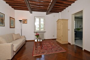 Um quarto com teto de madeira, radiador, sofá bege, tapete estampado, armário e porta que dá para uma cozinha.