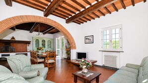 Una sala de estar con chimenea, muebles tapizados en verde, una zona de comedor con sillas y vistas al exterior a través de una ventana.