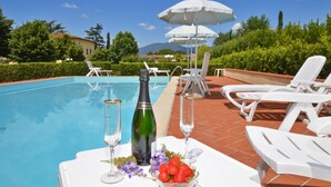 Zona de piscina con sillones, sombrillas y una mesa con una botella y vasos.