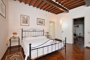 Quarto com estrutura de cama em metal, roupa de cama branca, piso de madeira e teto com vigas aparentes.