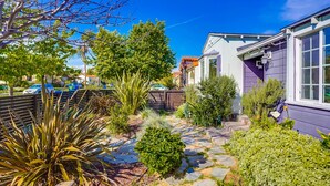 A residential garden with a stone pathway, various plants, and a house with purple siding.