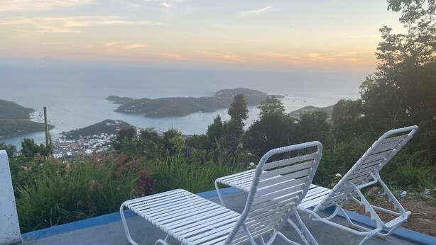 Deux fauteuils de relaxation en métal blanc sont placés sur une terrasse en béton, offrant une vue sur la mer et les îles lointaines.