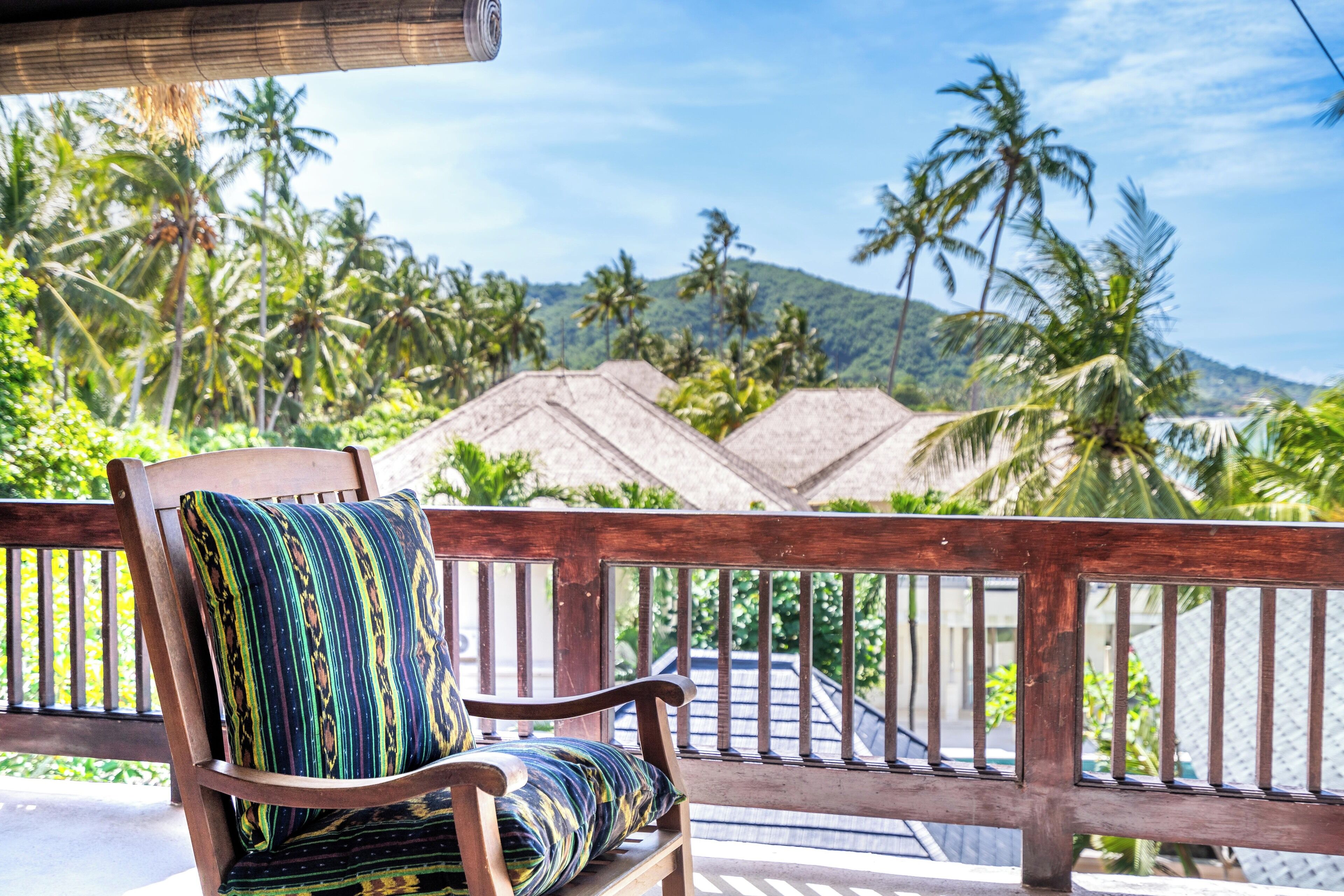 A wooden bench with a patterned cushion overlooking a tropical landscape with palm trees and a mountain in the distance.