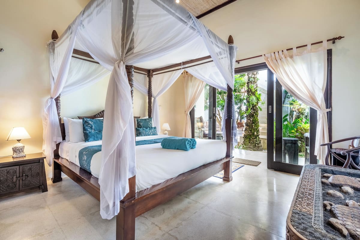 A four-poster canopy bed with white bedding, a nightstand with a lamp, and a view of the outdoors through a glass door.