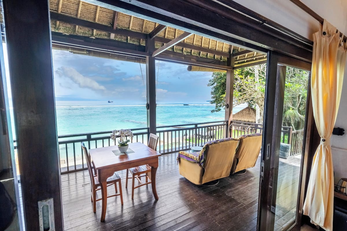A wooden deck with a table and chairs, a cushioned seating area, and a view of the ocean and boats.