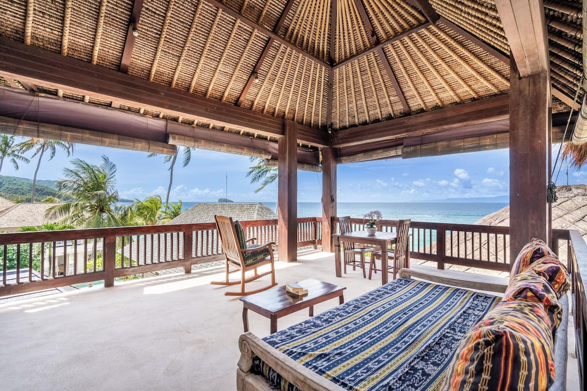 A covered outdoor seating area with a view of the ocean, wooden furniture, and a patterned bedspread.