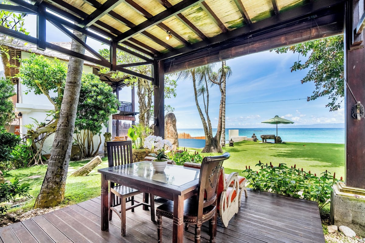 A wooden deck with a glass table and chairs overlooking a beach with a clear view of the ocean.