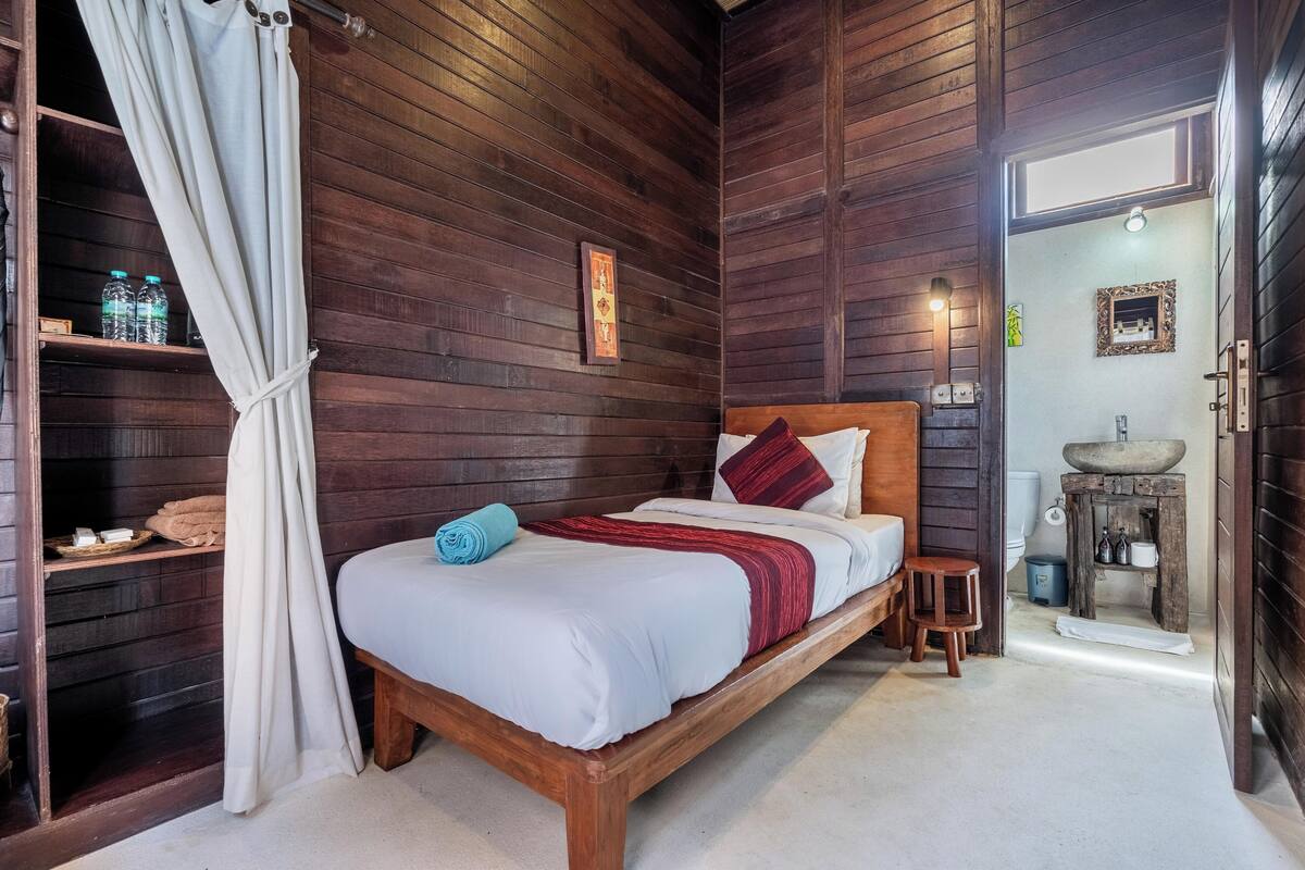 A wooden-paneled room with a bed, a small stool, a shelf with toiletries, and a door leading to a bathroom.