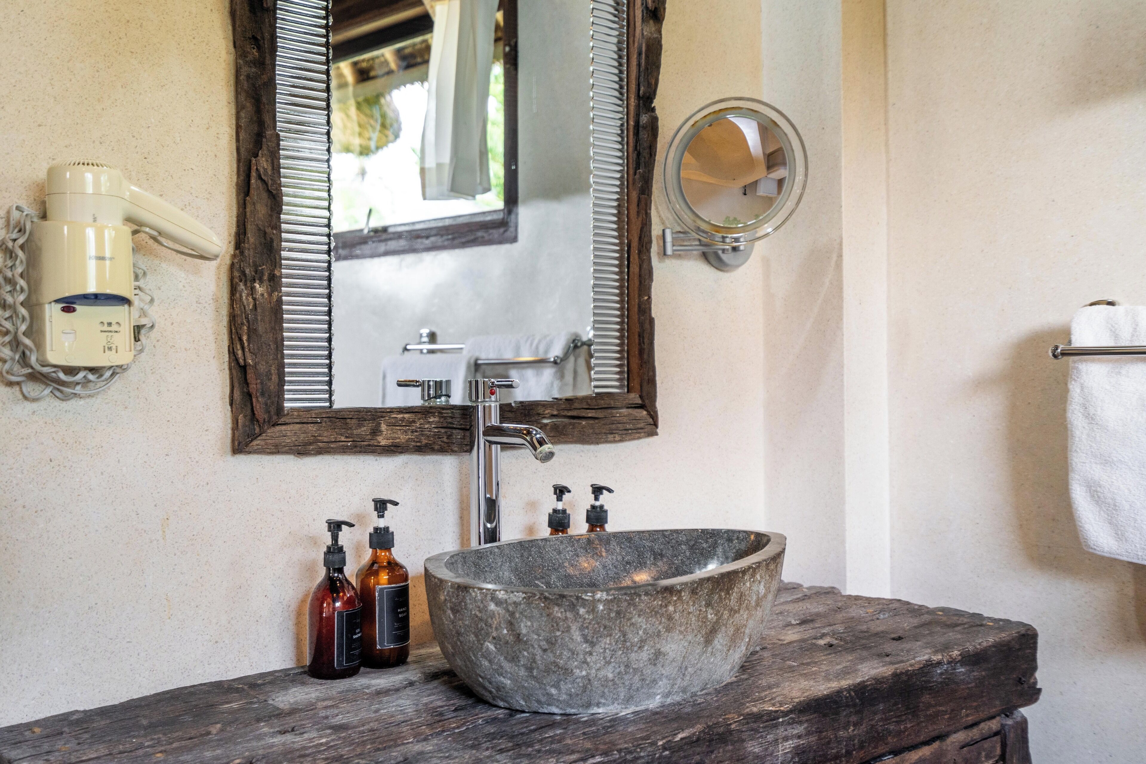 A bathroom with a stone sink, a wooden vanity, a mirror, a hairdryer, and two bottles of soap.