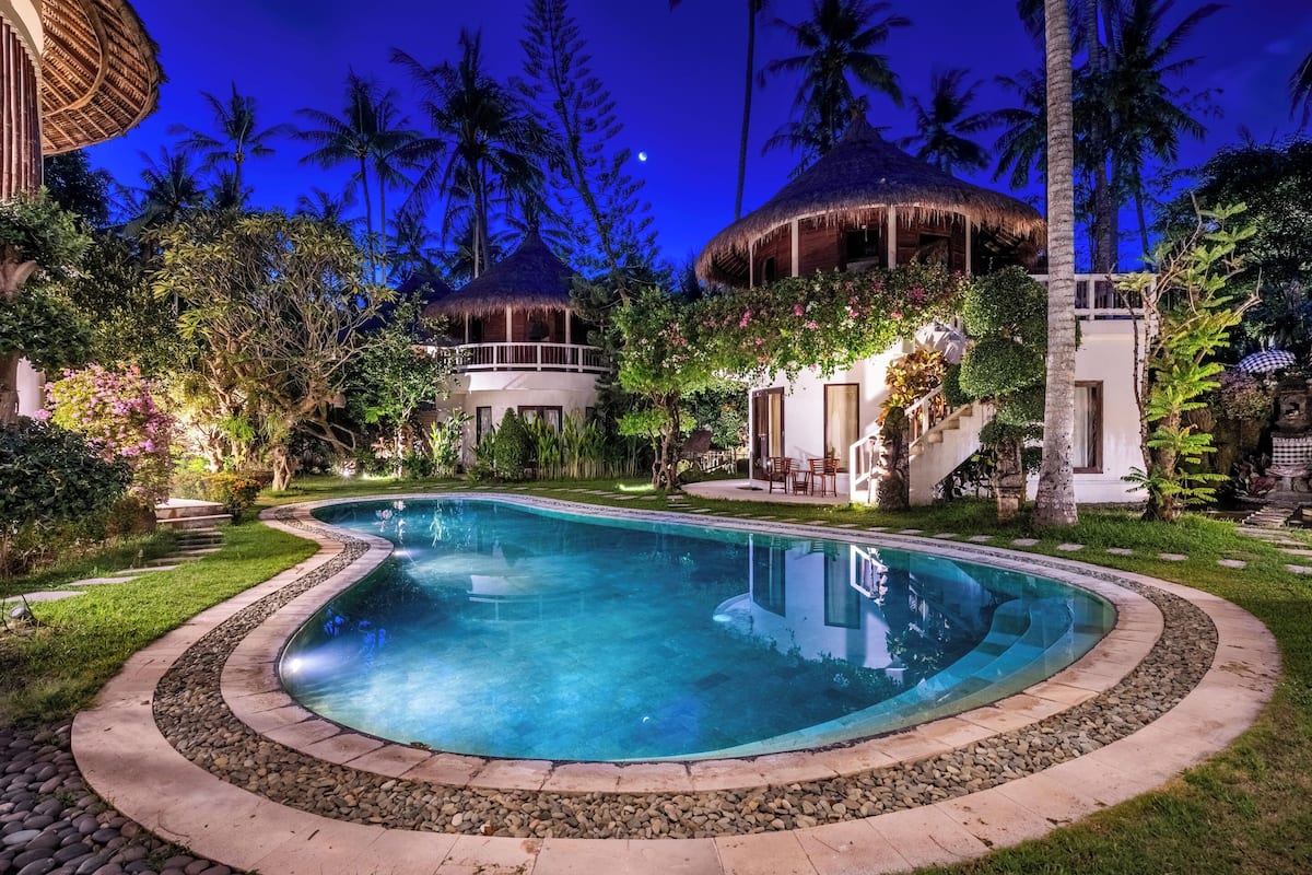A pool surrounded by lush greenery, a thatched-roof structure, and a white building with a balcony.
