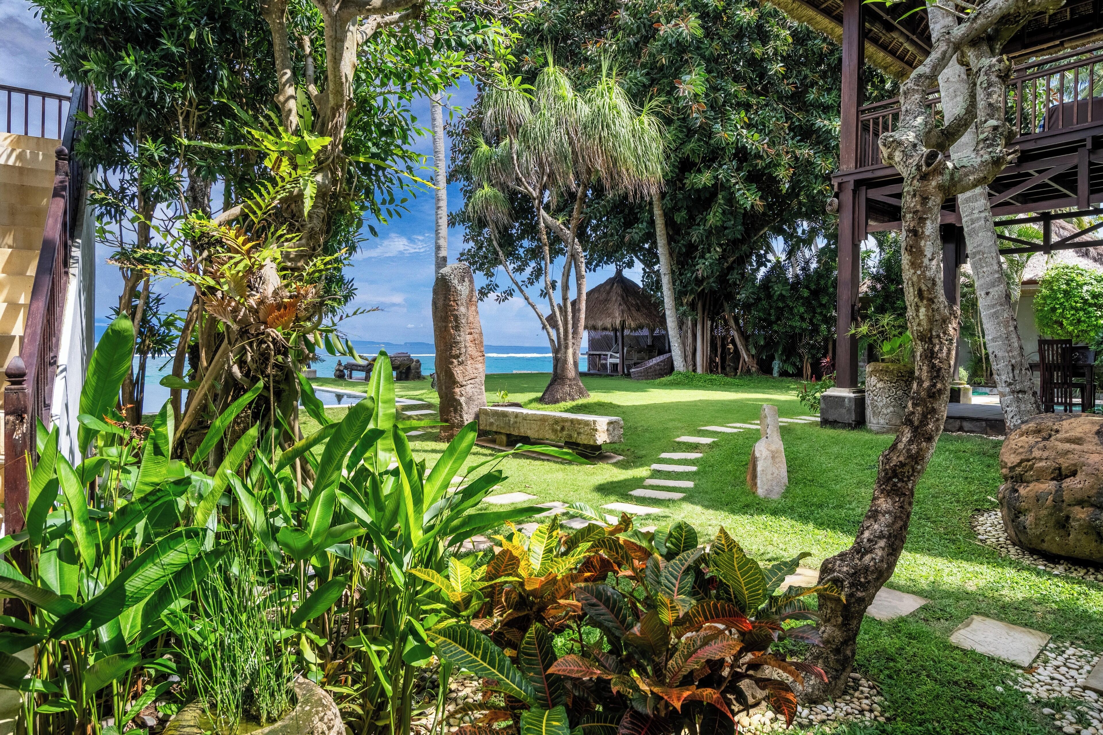 Un jardín tropical con una estructura de techo de paja, un camino de piedra y abundante vegetación.