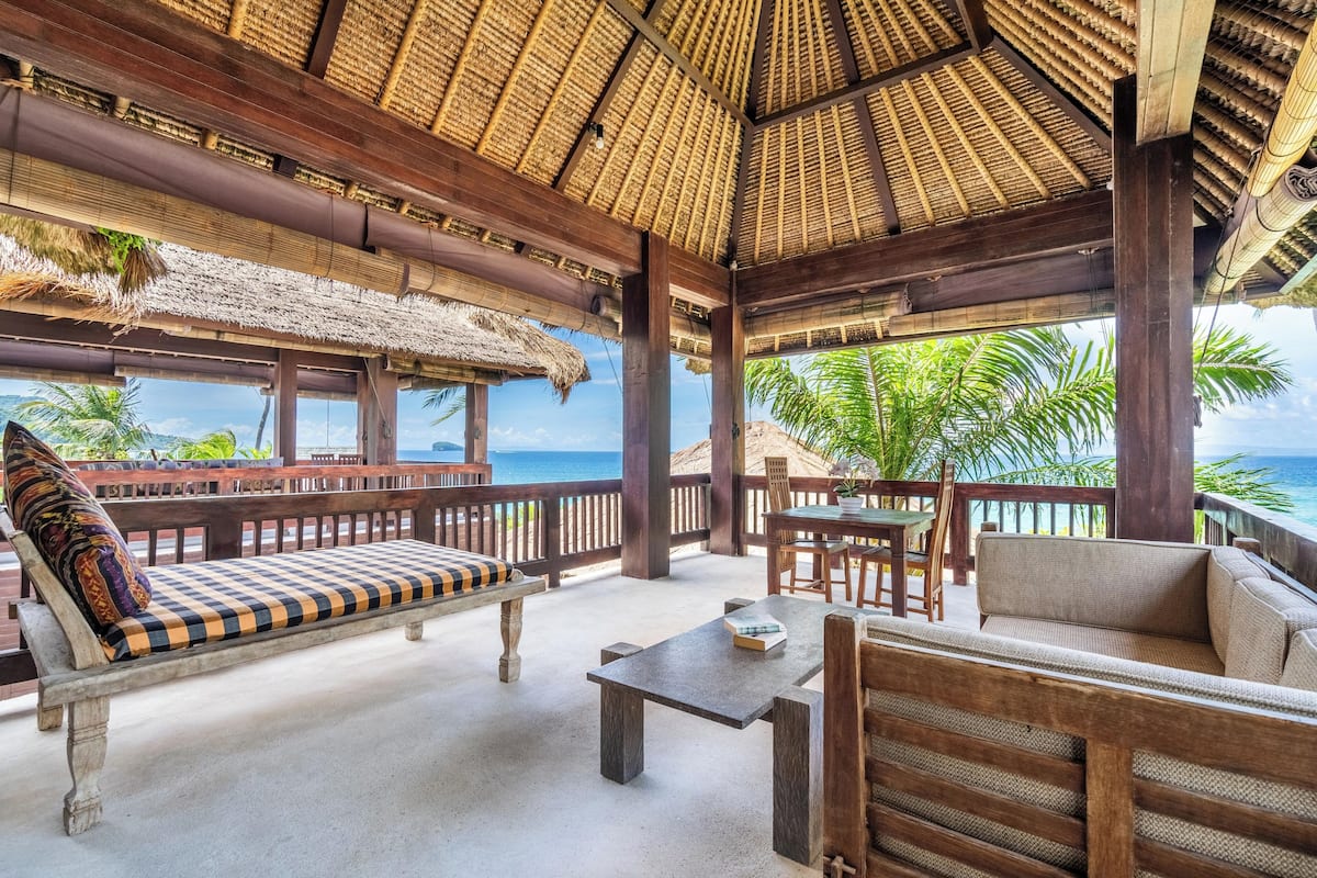 A covered outdoor seating area with wooden furniture and a thatched roof, overlooking a beach.