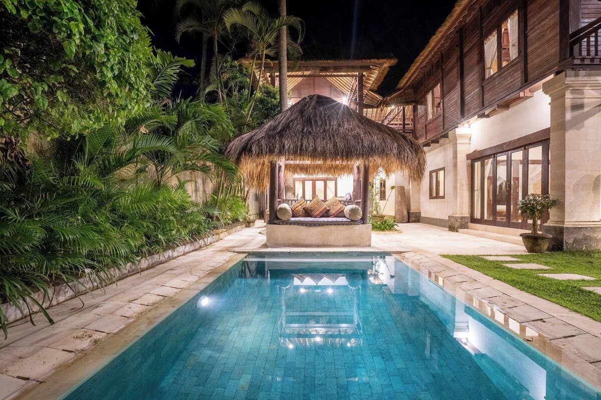 A swimming pool with a thatched-roof structure and a bench, surrounded by lush greenery and a well-lit building.