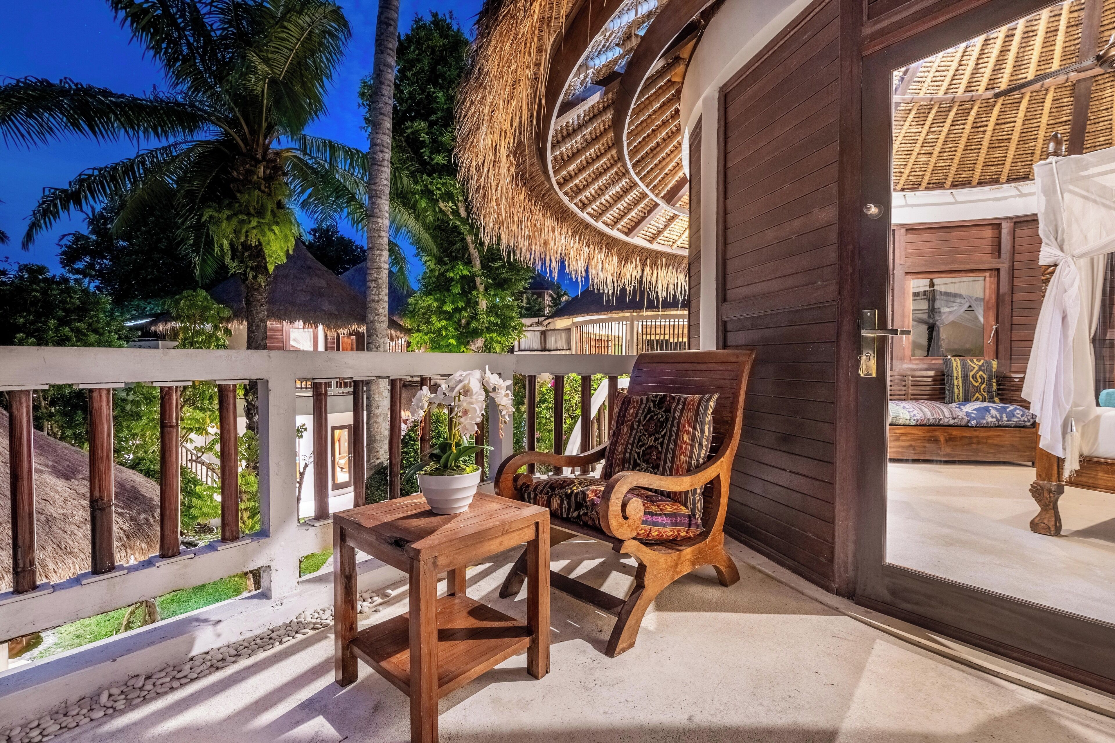 A wooden deck with a table and chair, a thatched roof structure, and a view of palm trees and other buildings.