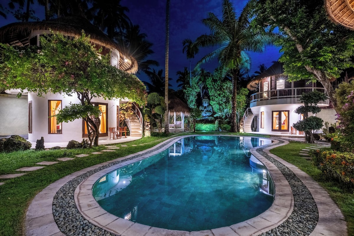 A swimming pool surrounded by lush greenery and two white buildings with thatched roofs.