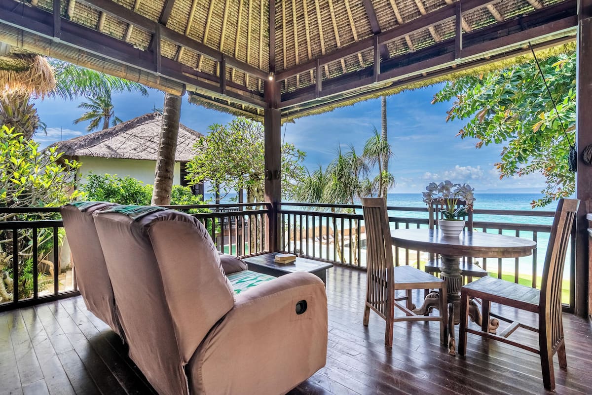 A covered outdoor seating area with a view of the ocean, a table with chairs, and a thatched roof structure.