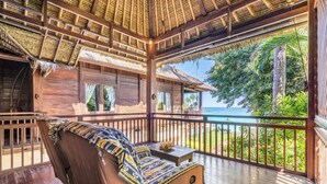 A wooden deck with a sofa and a table, overlooking a tropical beach and ocean. - The Village of Angels (Manggis)