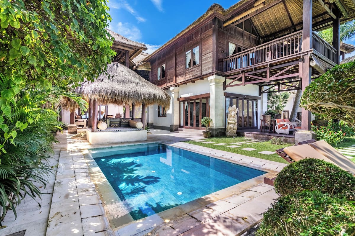 A pool area with a thatched-roof structure, a wooden deck, and a large house with balconies.