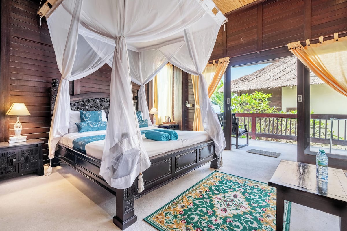 A bedroom with a four-poster bed, a nightstand, a lamp, and a view of the outdoors.