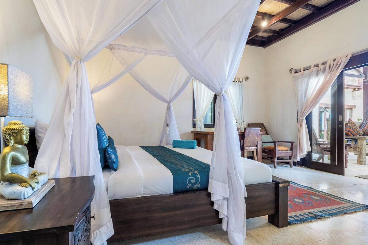 A room with a four-poster bed, a Buddha statue, and a view of the outside.