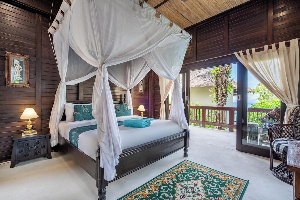 A four-poster bed with a canopy, a nightstand with a lamp, a patterned rug, and a view of the outdoors through a glass door.